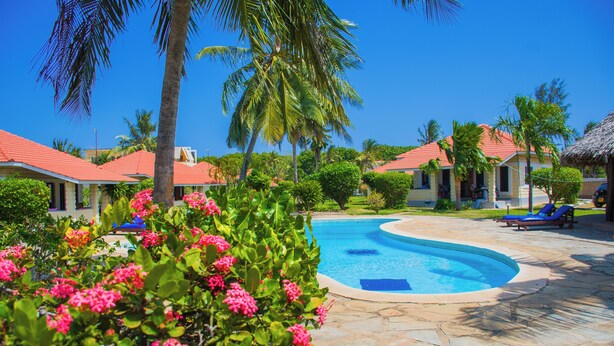 A swimming pool surrounded by stone tiles, palm trees, and houses with red roofs. - Doric Cottages Diani (Diani Beach)