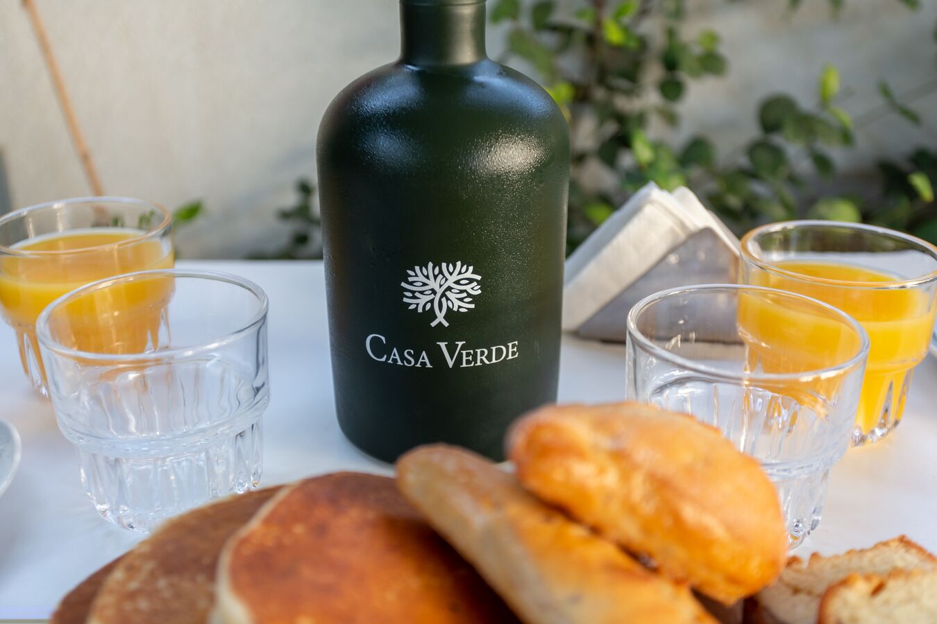 Una botella con la etiqueta "Casa Verde", vasos de jugo de naranja y pan sobre una mesa.