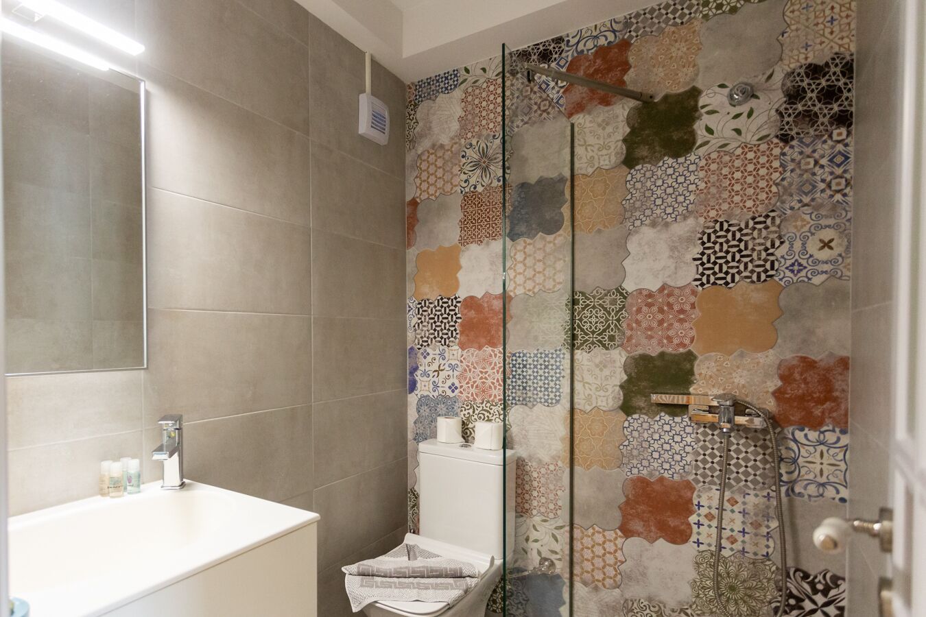 Un baño con pared de ducha enlosada, un lavamanos con espejo y un inodoro.