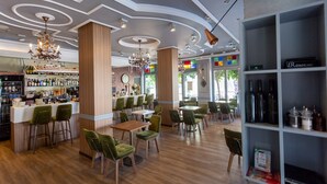 L’intérieur d’un restaurant moderne présente un bar, des chaises à revêtement vert, des tables en bois et une variété de bouteilles exposées sur des étagères.
