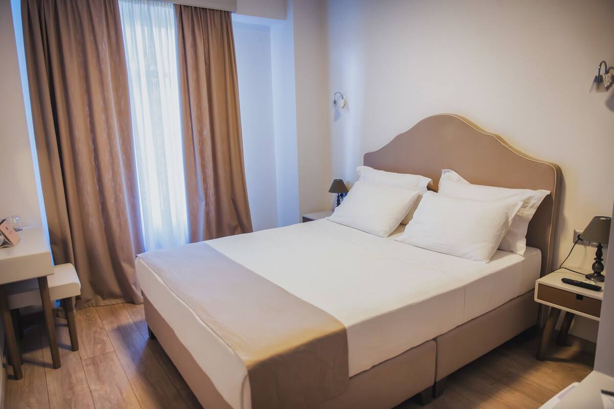 A neatly made bed with white linens, a bedside table with a lamp, and a desk with a chair.