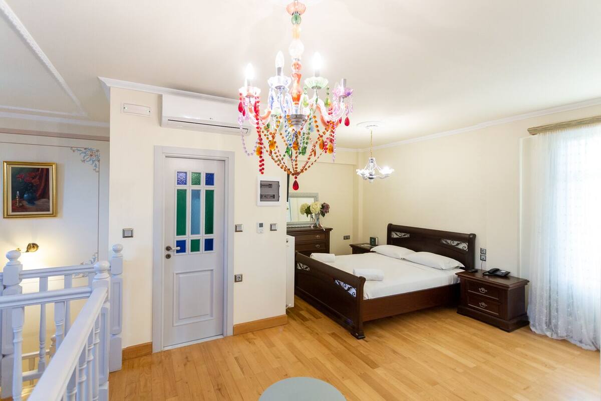 A bedroom with a wooden floor, a bed with white linens, a chandelier, a bedside table, a nightstand, a door with frosted glass panels, and a painting on the wall.