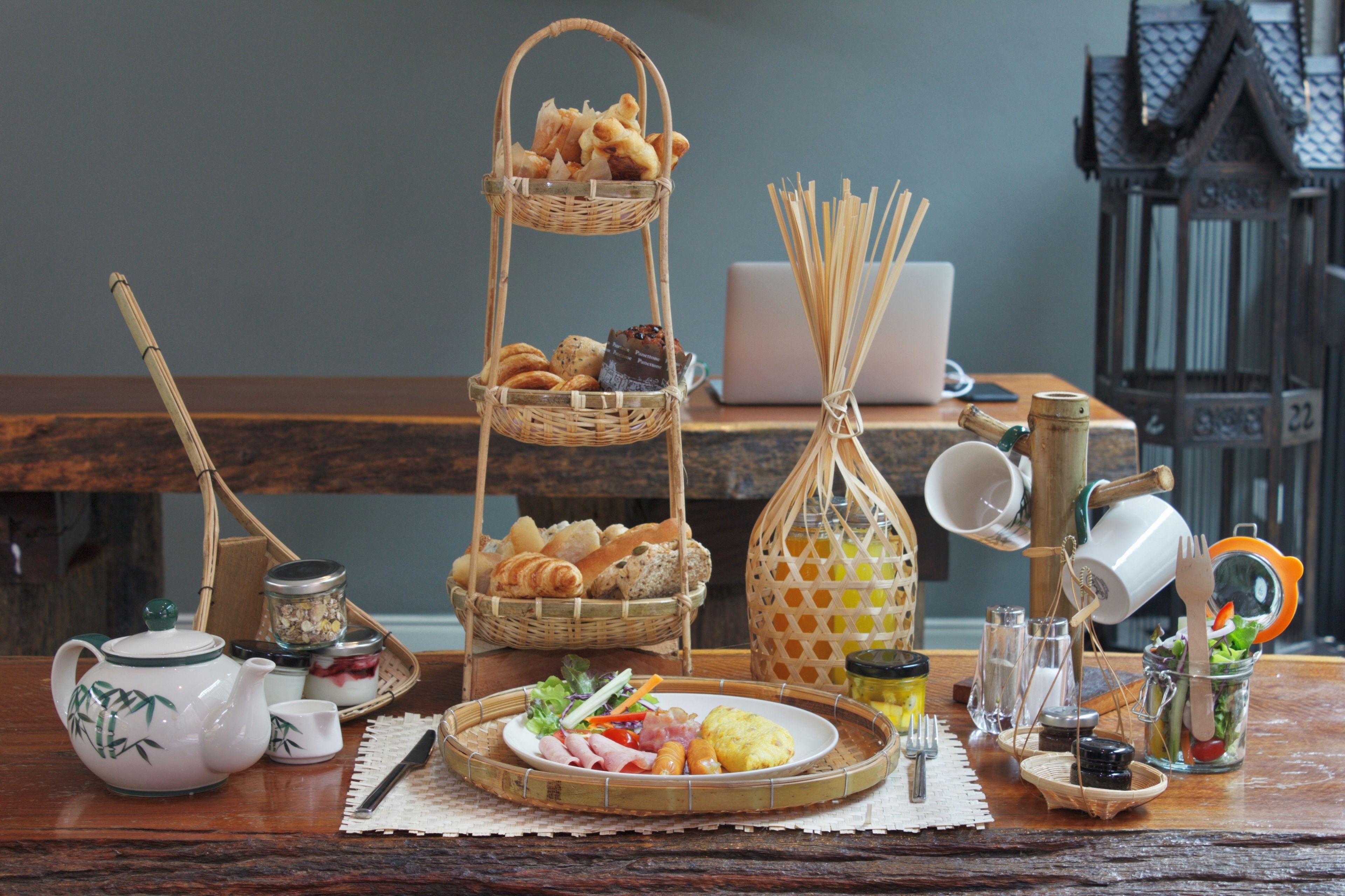A table set with a teapot, cups, a laptop, a basket of bread, a bottle, and a glass jar with a salad.
