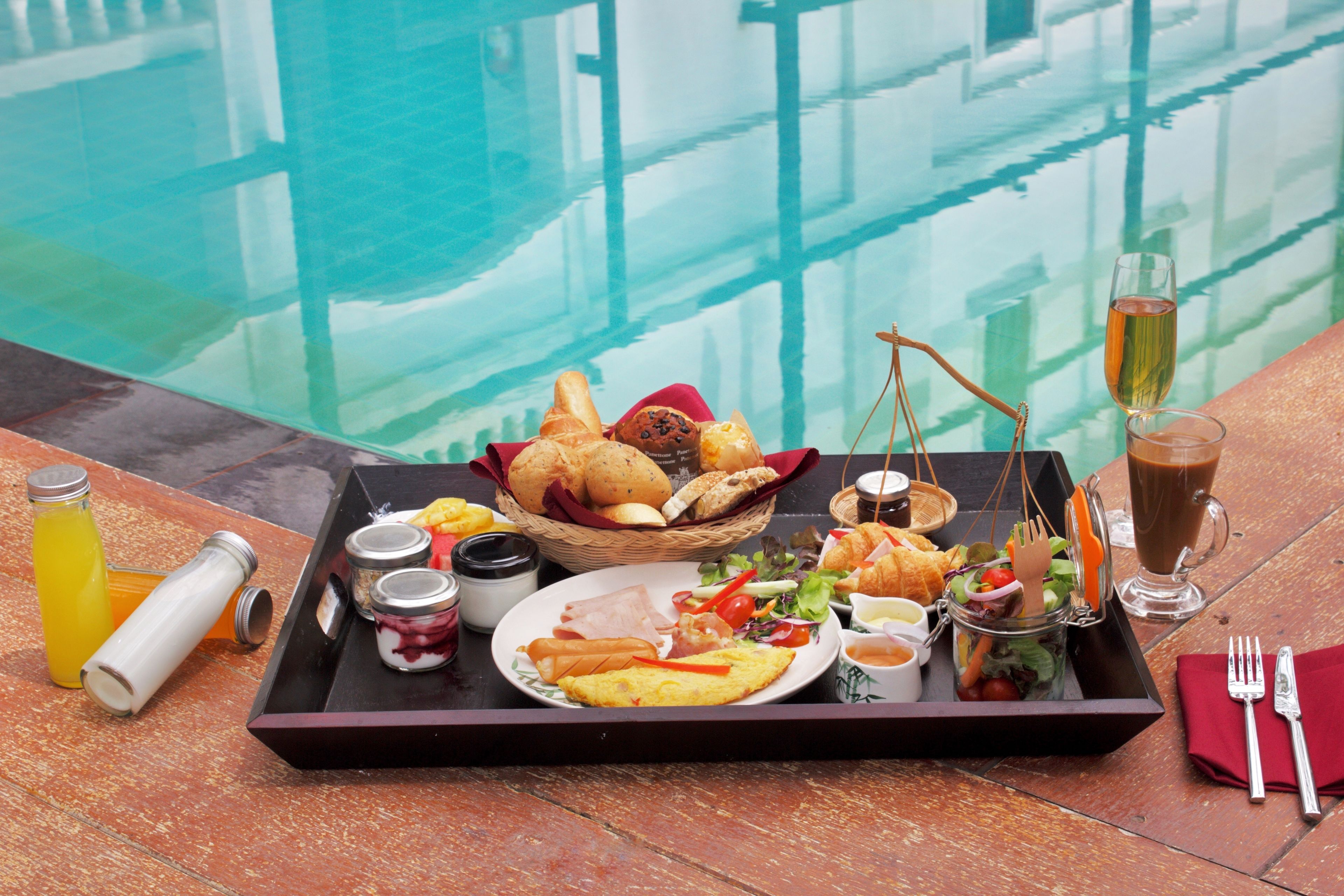 A tray with a variety of breakfast items including eggs, sausages, and toast, placed on a wooden table by a pool.