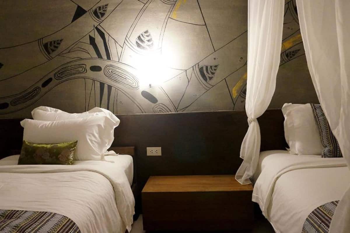 A hotel room with two beds, each with white bedding and a green pillow. The room features a modern abstract wall design and a wooden nightstand between the beds.
