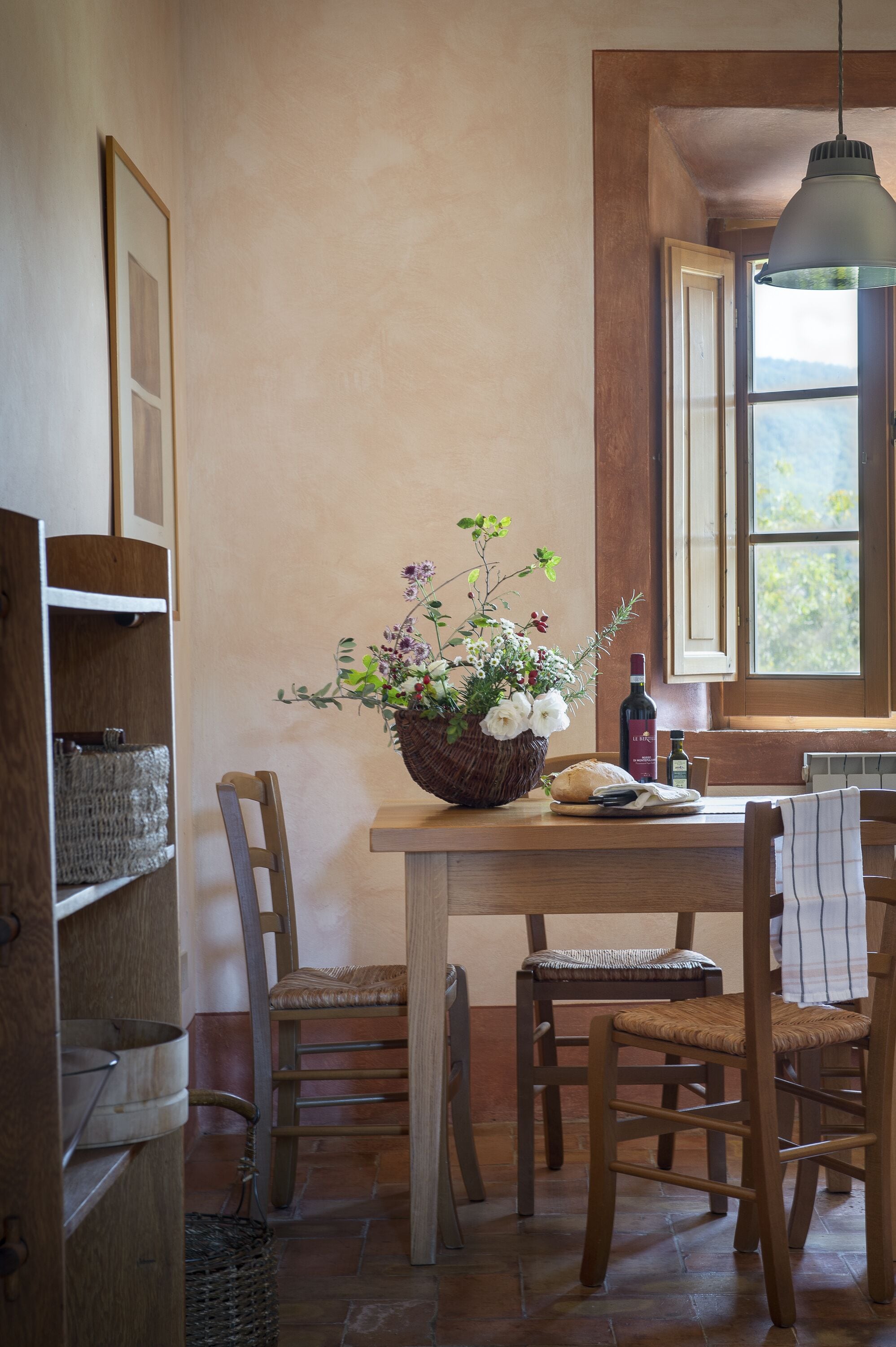 A dining area with a wooden table, wicker chairs, a wicker basket with flowers, a bottle of wine, and a window with a view of the outdoors.