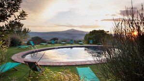 A circular pool with lounge chairs around it, surrounded by greenery and a mountain in the background. - Azienda Agricola Chiarentana (Chianciano Terme)