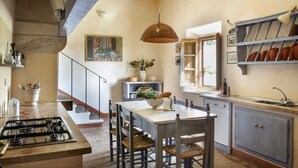 A kitchen with a gas stove, wooden chairs, a hanging light fixture, and a window overlooking the garden. - Azienda Agricola Chiarentana (Chianciano Terme)