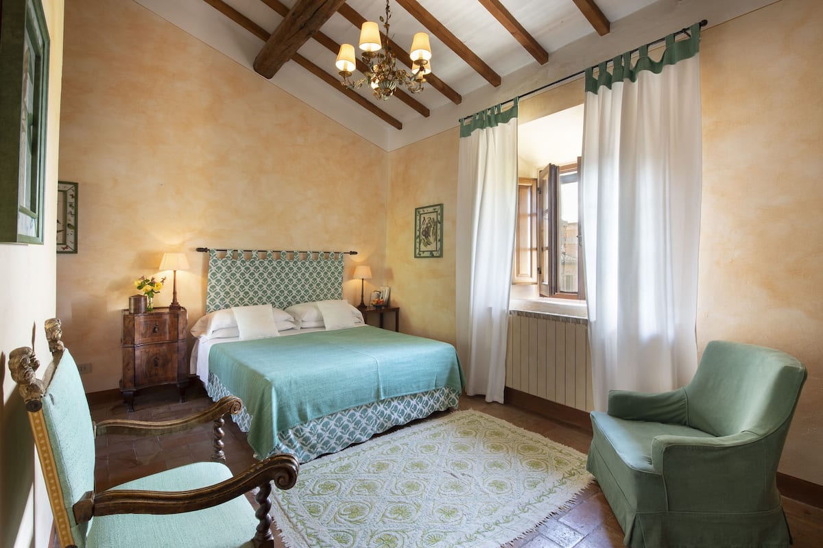 Une chambre à coucher avec un lit, deux tables de chevet, une chaise, un lustre et une fenêtre avec des rideaux.