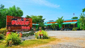 A hotel entrance with a large sign, a gravel driveway, and colorful buildings in the background. - Kwanjai Resort (Tha Mai)