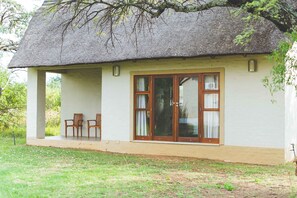 A thatched-roof cottage with a covered porch, two wooden chairs, and a window with curtains. - Deruxa Cottages (De Wagensdrift)