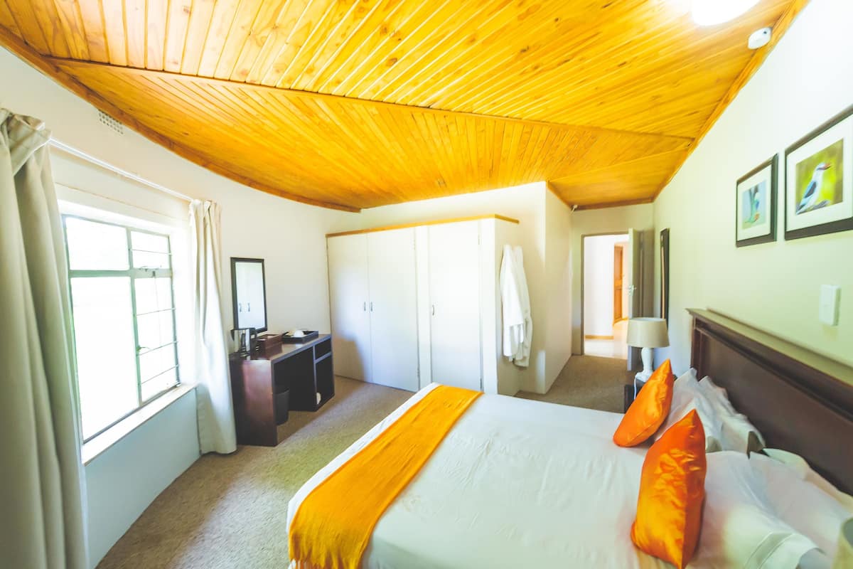 A hotel room with a wooden ceiling, a bed with white and orange bedding, a desk with a chair, and a wardrobe.