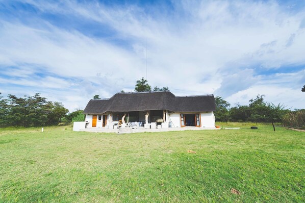 A house with a thatched roof, surrounded by a grassy field and trees. - Deruxa Cottages (De Wagensdrift)