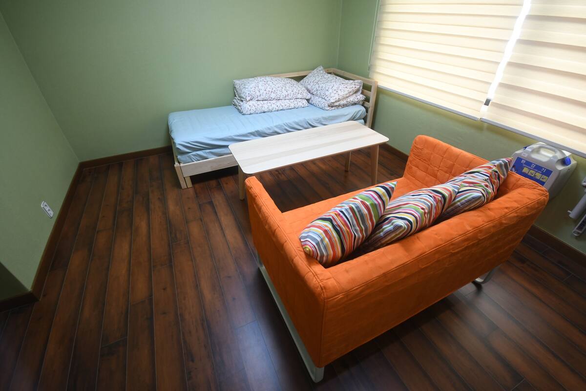 A compact room with a bed, a small table, an orange sofa, and a window with blinds.