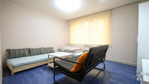 A small, sparsely furnished room with a bed, a sofa, a chair, and a window with blinds. - BLUEHUM Guesthouse - Hostel (Incheon)