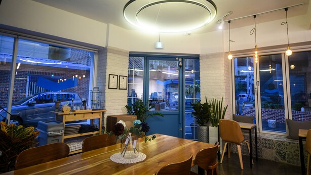 A modern dining area with a wooden table, chairs, and a vase of flowers. There are large windows with views of the street and a parked car outside. - BLUEHUM Guesthouse - Hostel (Incheon)