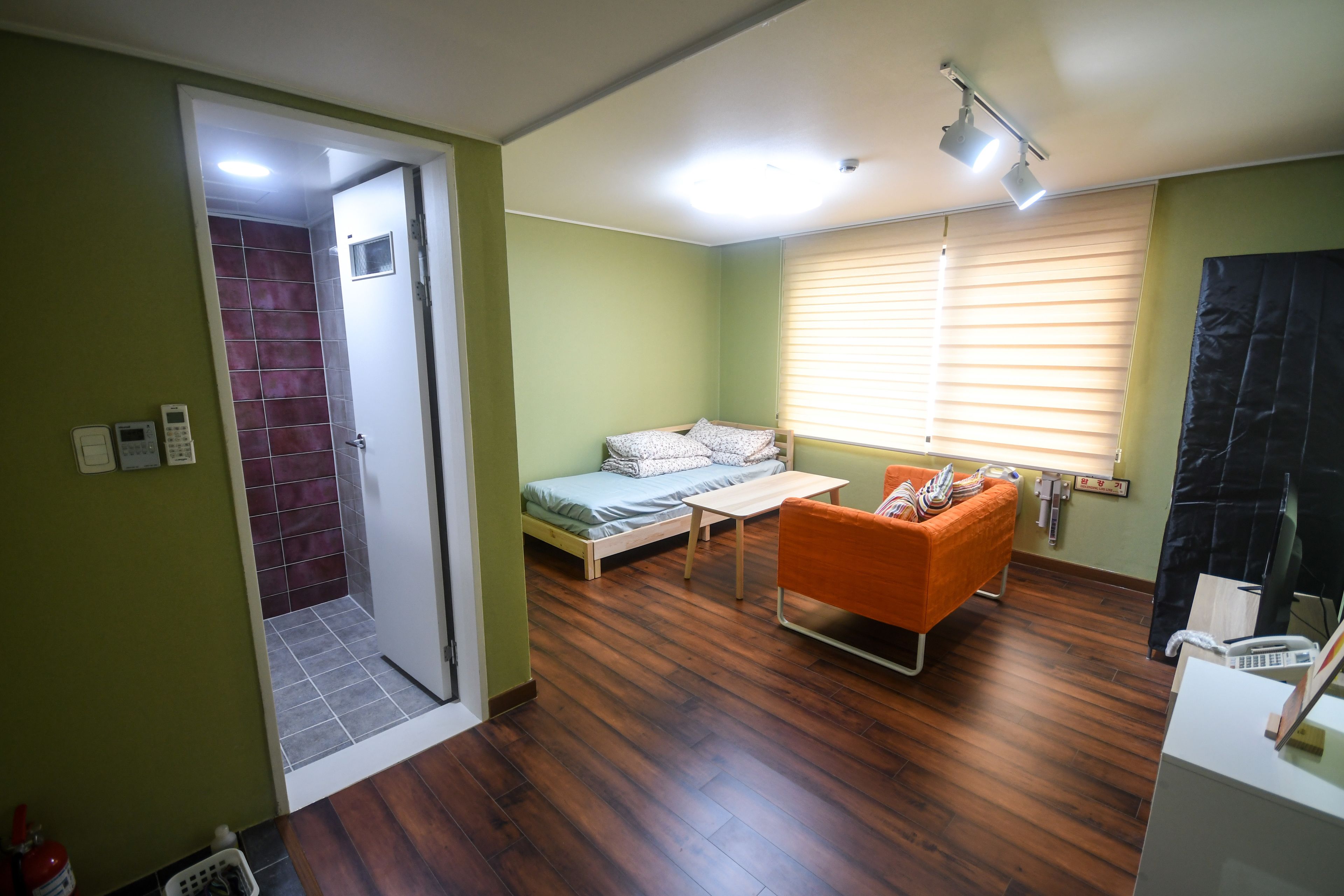 A compact room with a bed, a small table, an orange chair, a window with blinds, and a door leading to a bathroom.