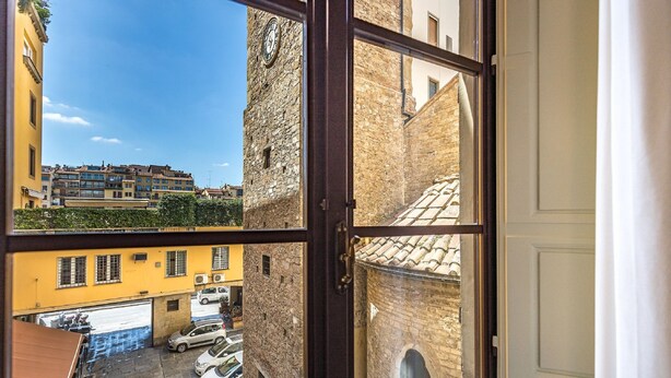 A view through a window of a narrow street with parked cars and a building with a clock tower. - Residenza Alessandra (Florence)