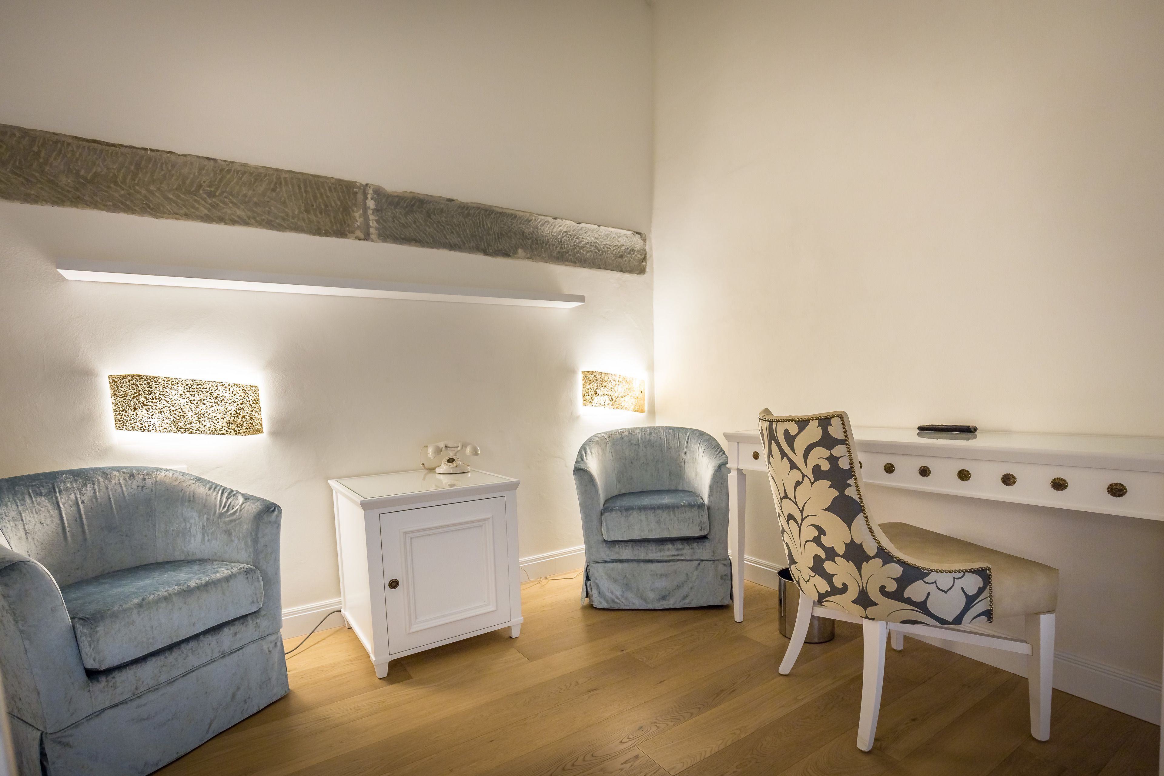 Ein Zimmer mit einem weißen Schrank, zwei blauen Sesseln, einem gemusterten Sessel und einem Schreibtisch mit einer Lampe.