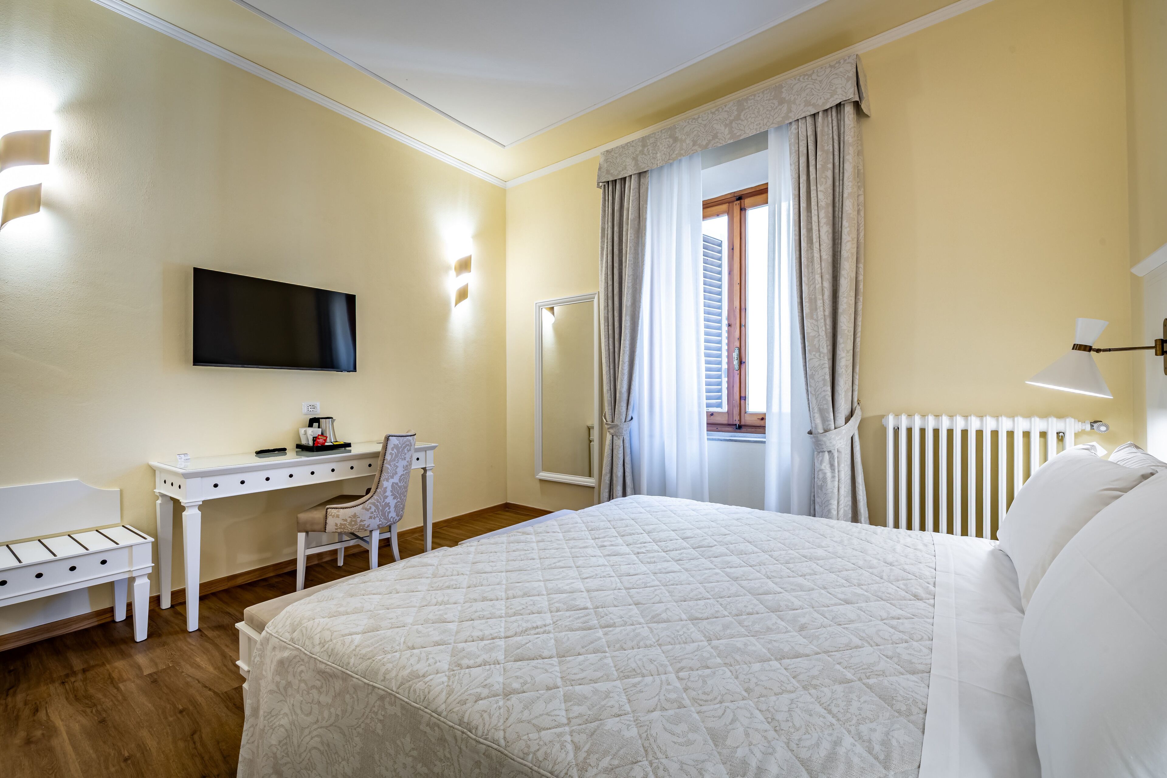 Una camera d'albergo con un letto, una scrivania, una sedia, una televisione e una finestra con tende.