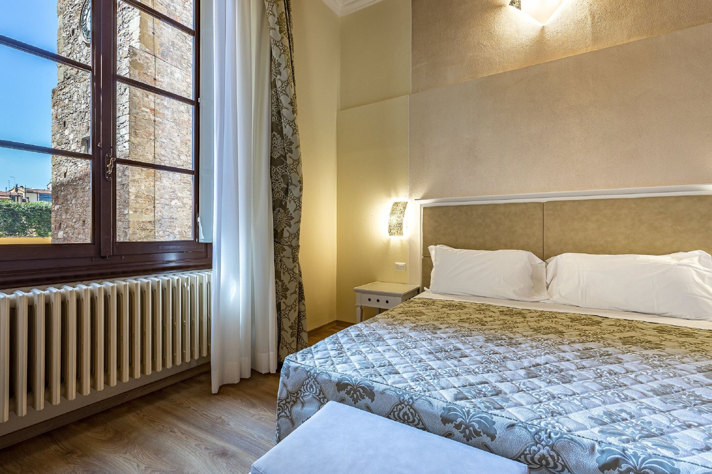 Ein Hotelzimmer mit großem Fenster, Heizkörper, Nachttisch mit Lampe und einem Bett mit gemustertem Überwurf.