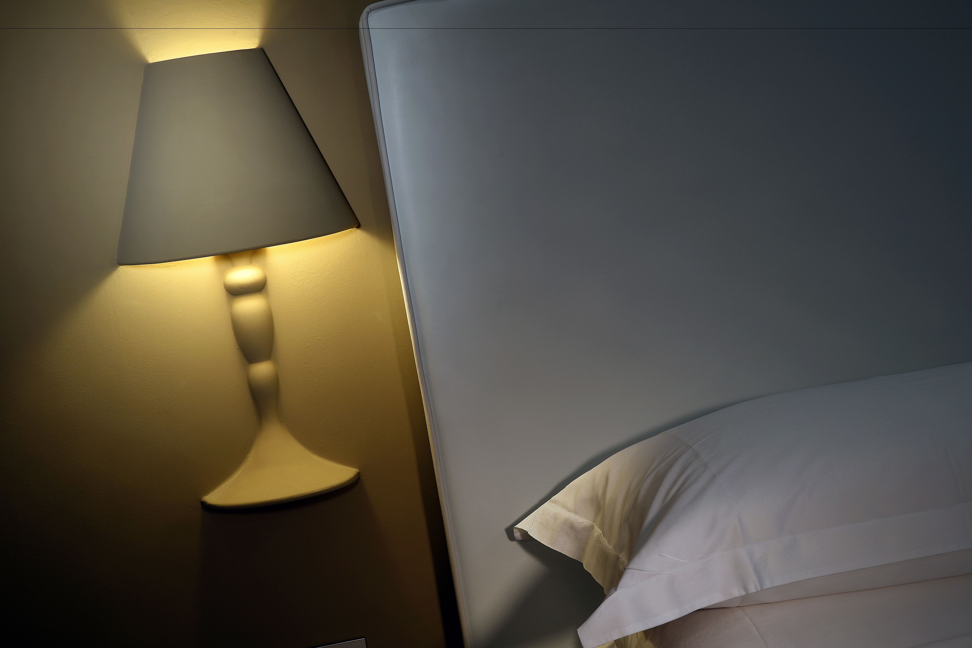 Una cama con ropa blanca y una lámpara fijada a la pared.