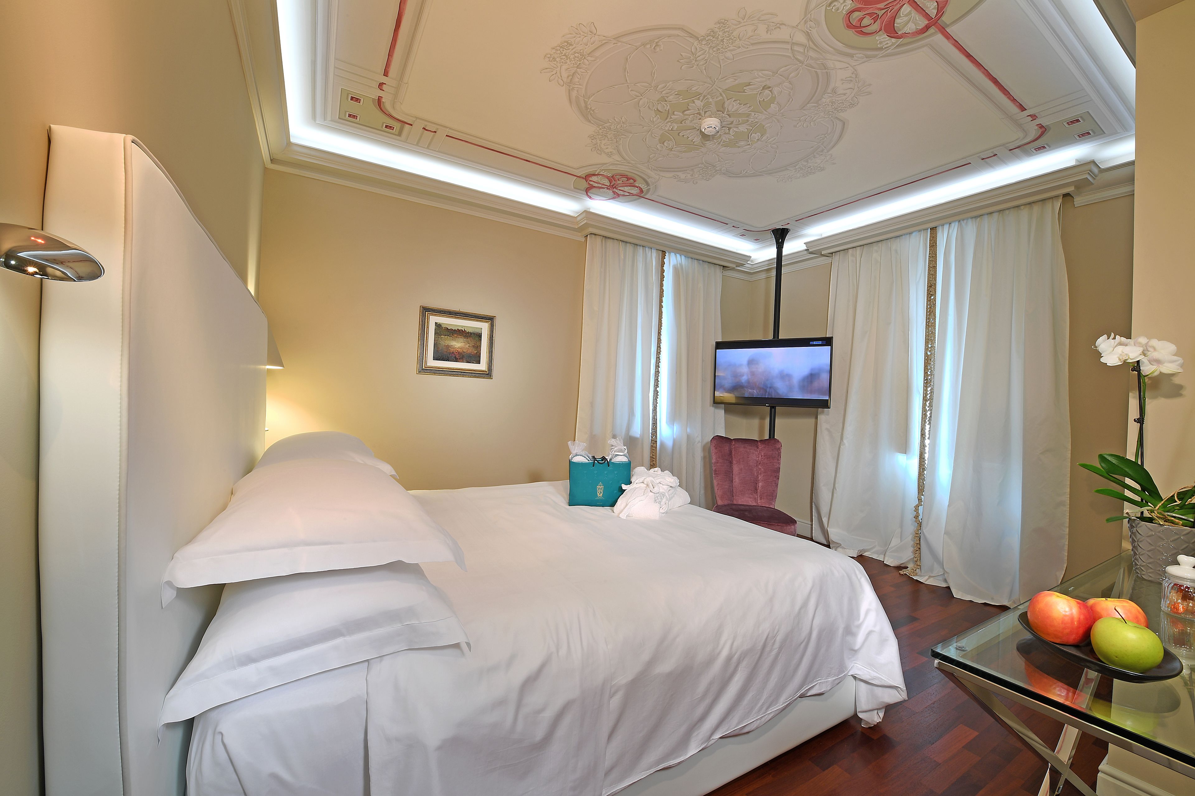 A hotel room with a bed, a TV, a glass table with fruits, and a potted plant.
