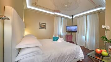 A hotel room with a bed, a TV, a glass table with fruits, and a potted plant.