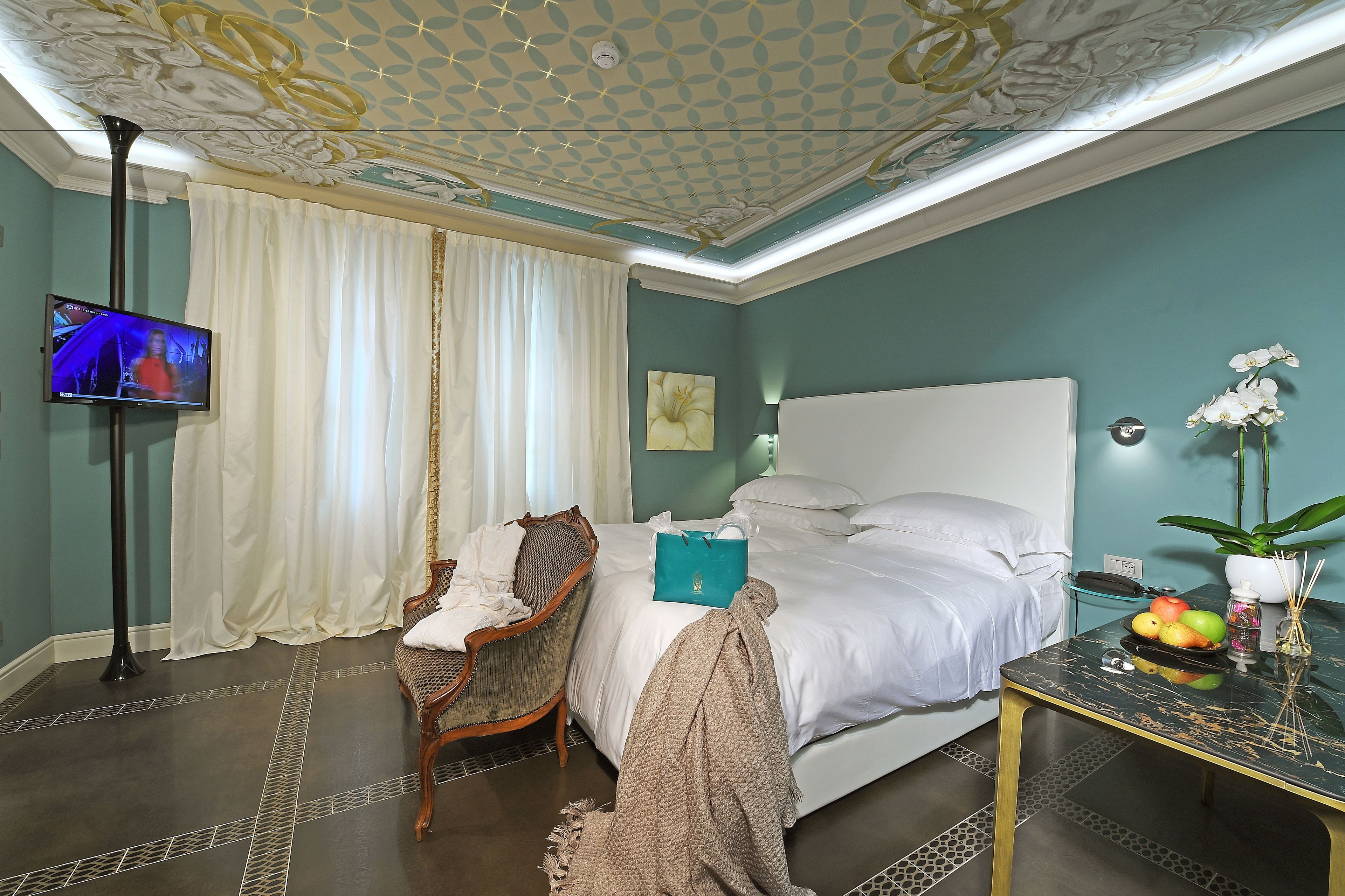 A hotel room with a bed, a television, a chair, a table with fruit, and a plant.