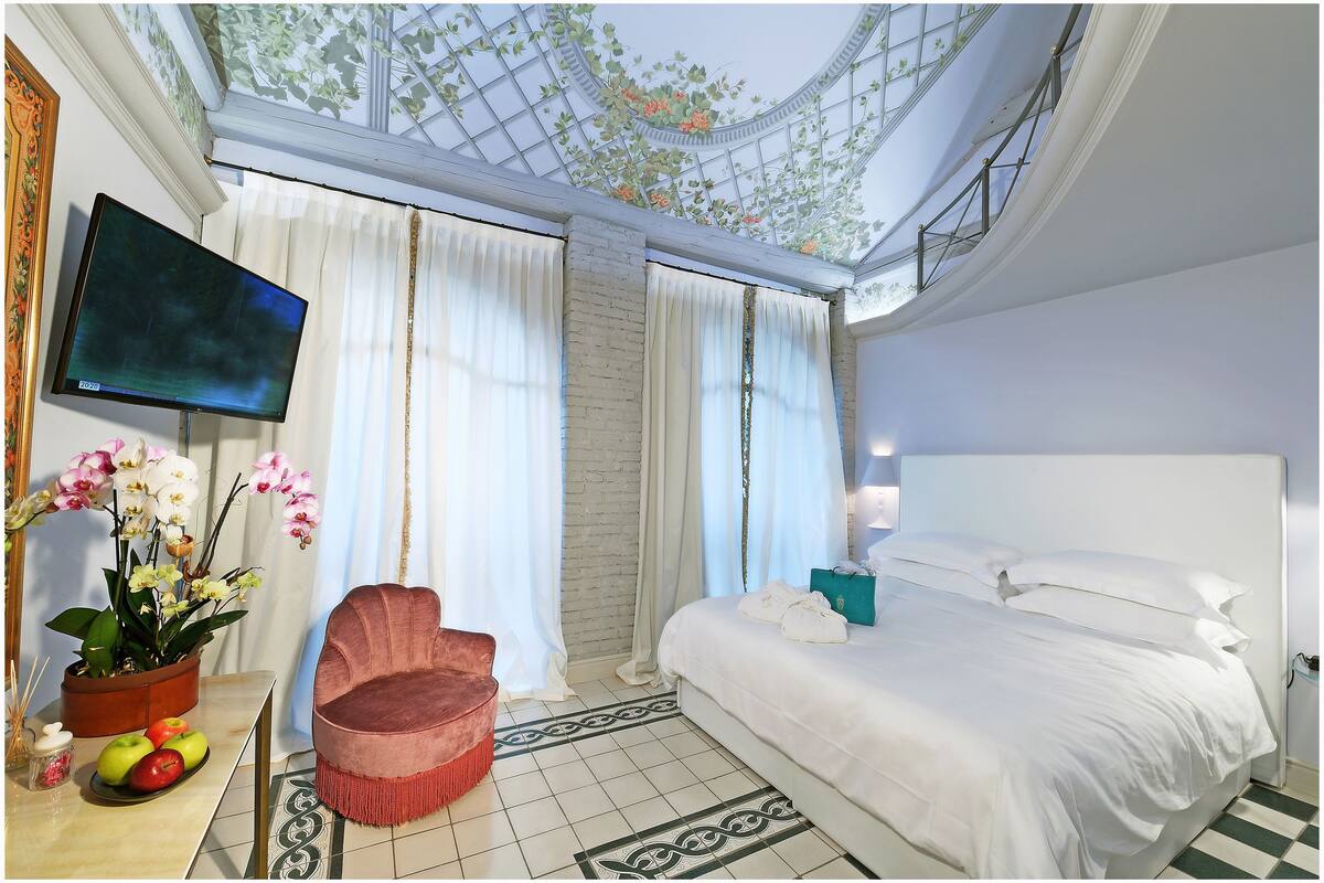 A hotel room with a bed, a TV, a pink chair, a plant, and a fruit bowl.