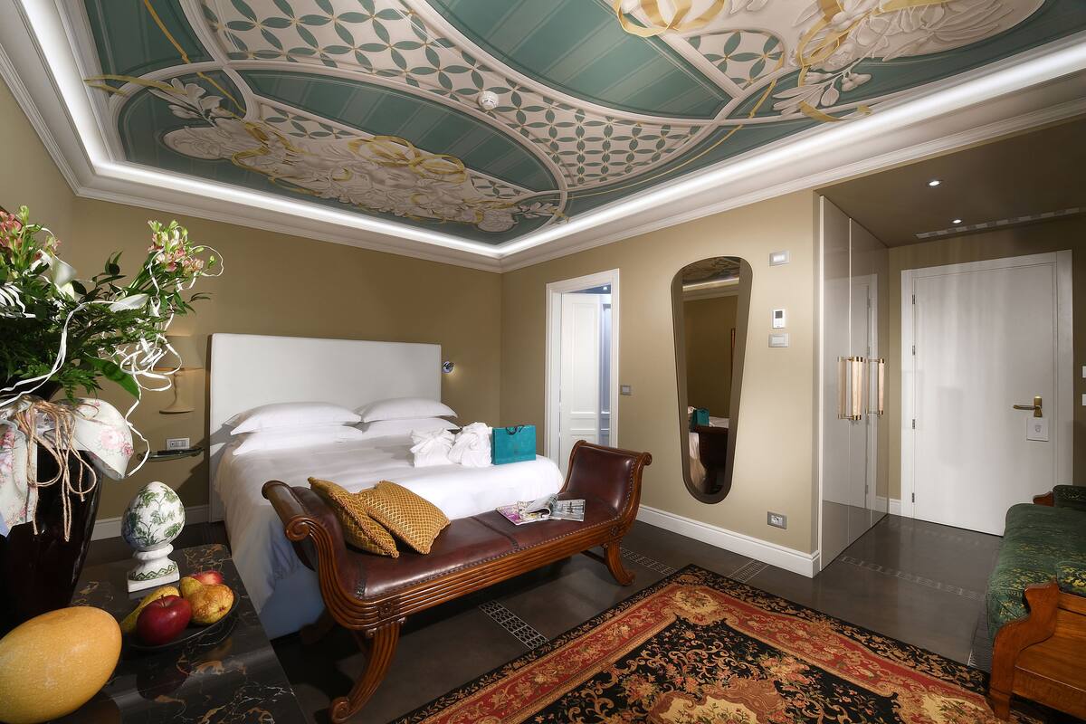 A bedroom with a bed, a bench, a large mirror, and a door leading to another room.