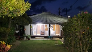 Une maison traditionnelle de style japonais, avec un toit en tuiles, une terrasse en bois et un jardin agrémenté de plantes en pot et d’un sentier en pierre.
