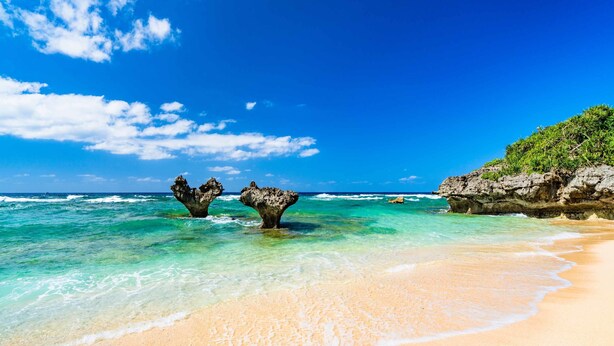 Une plage à l’eau turquoise et limpide, bordée d’un rivage sablonneux et ponctuée de formations rocheuses.