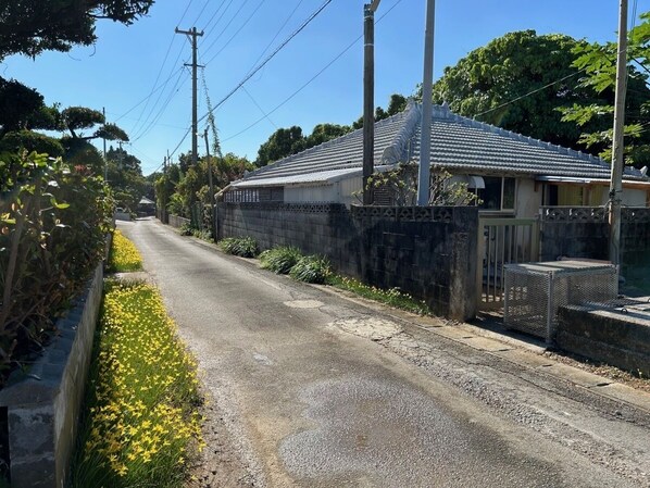 A residential street with a house featuring a tiled roof and a fenced yard with yellow flowers.