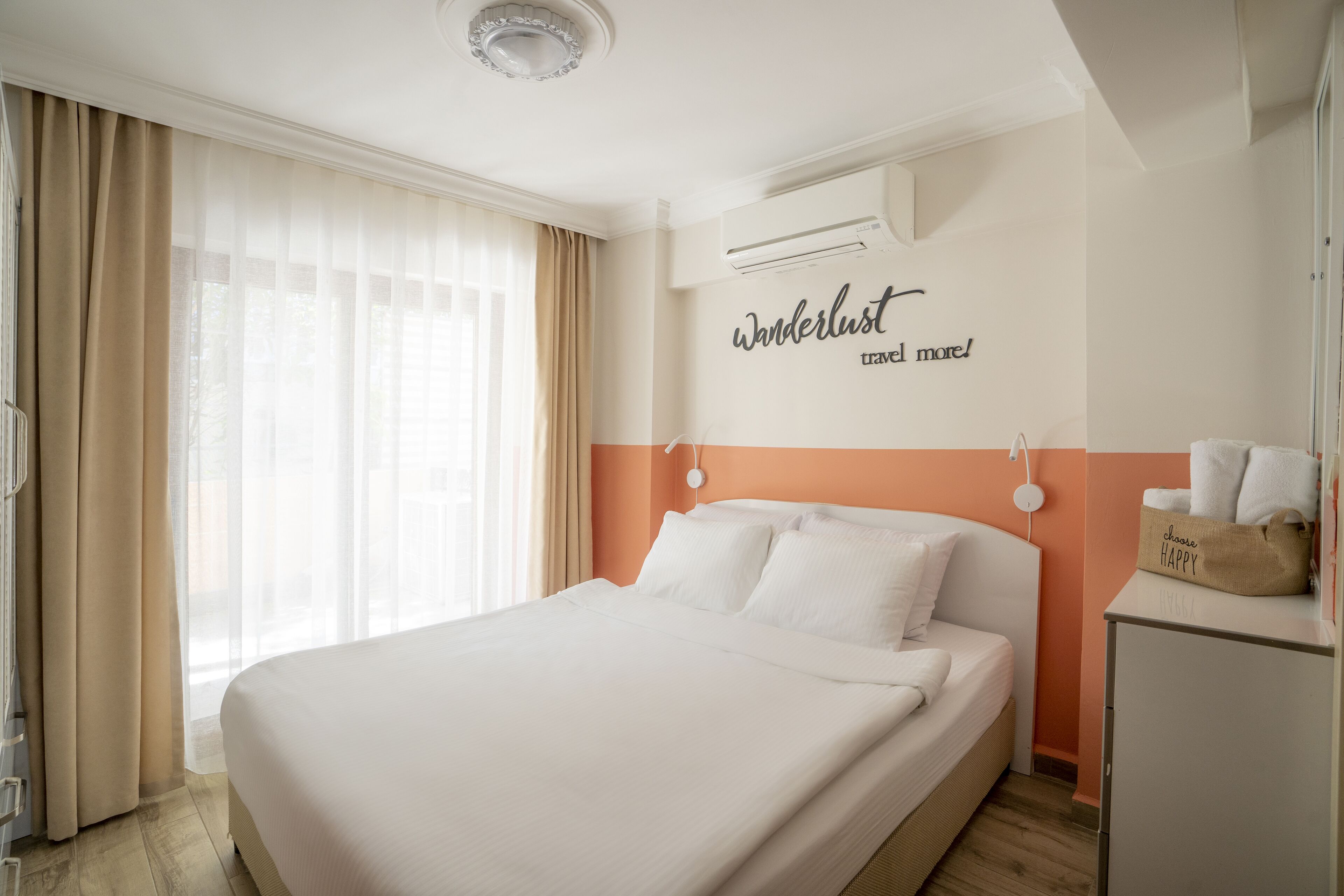 A hotel room with a large bed, beige curtains, an air conditioning unit, and a wall with a 'wanderlust' sign.