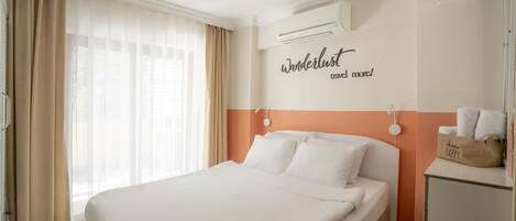 A hotel room with a large bed, beige curtains, an air conditioning unit, and a wall with a 'wanderlust' sign.