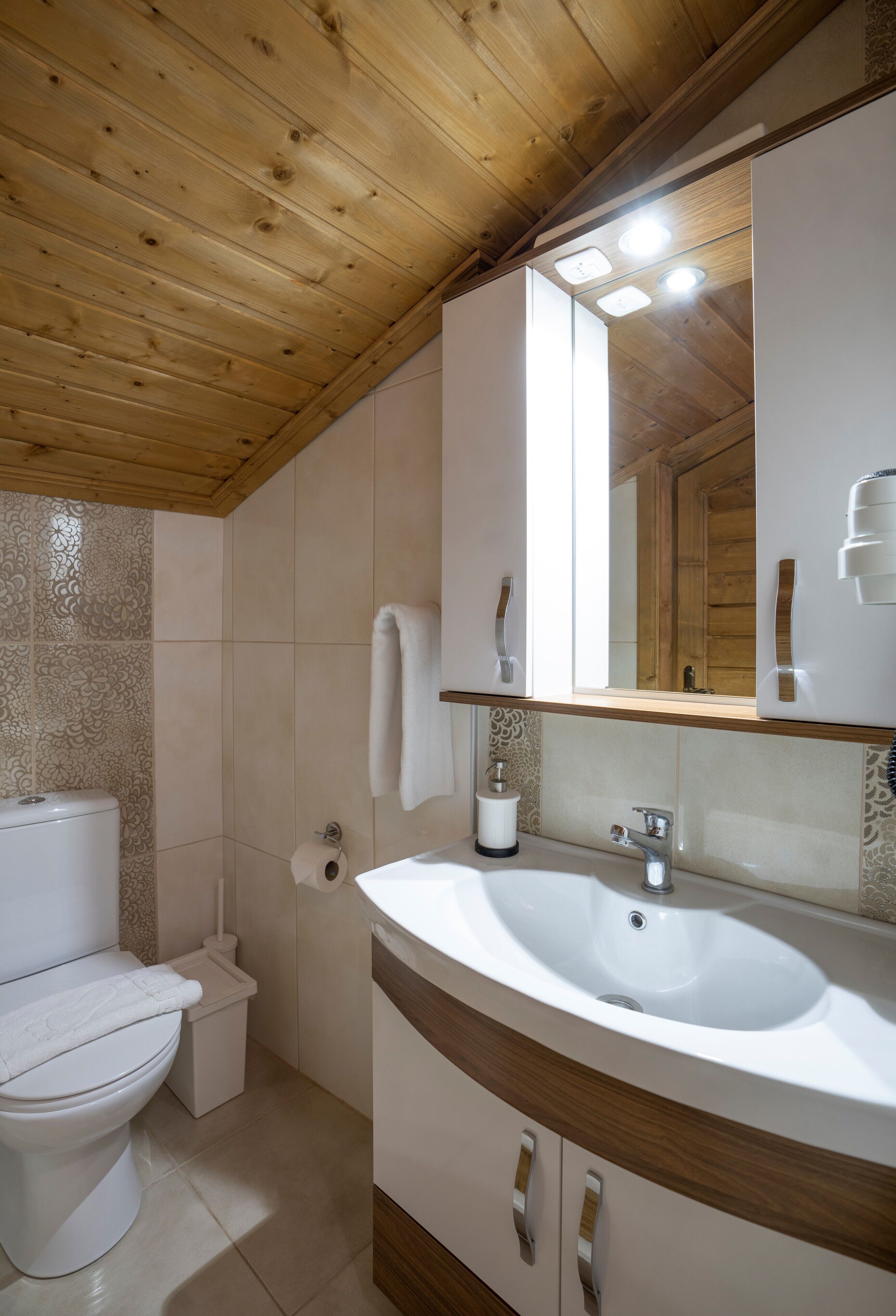 Un baño con techo de madera, inodoro blanco, lavamanos con espejo y azulejos estampados en las paredes.