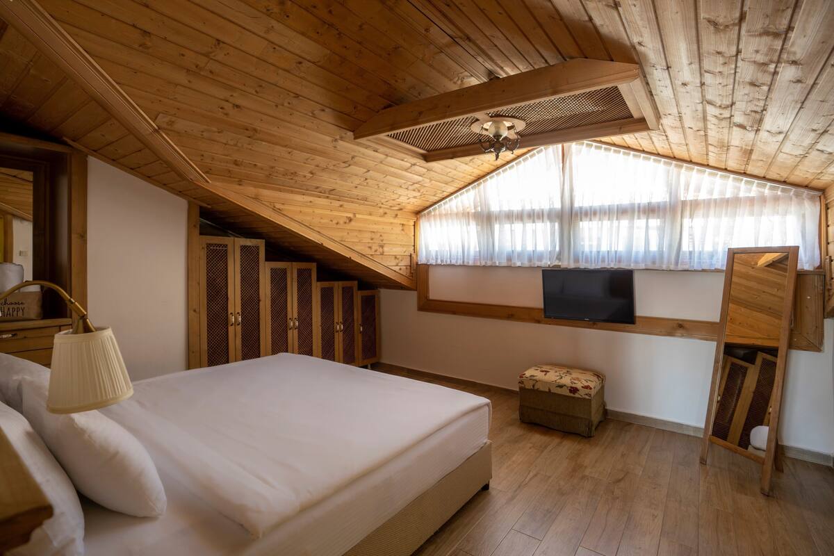 A bedroom with a wooden ceiling, a bed, a window with curtains, a television, and a small bench.