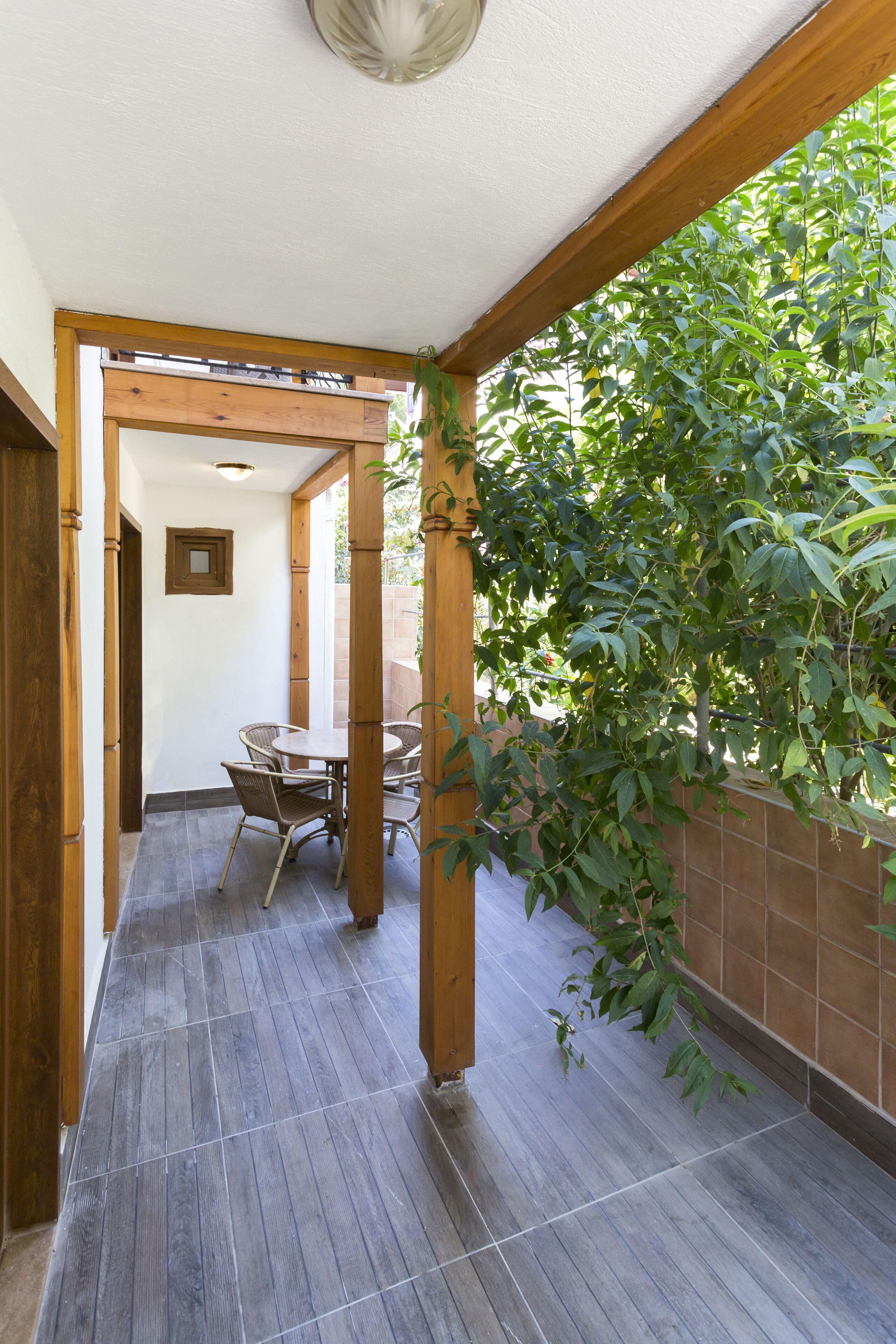 A covered patio area with a wooden table and chairs, a tiled floor, and a view of greenery outside.
