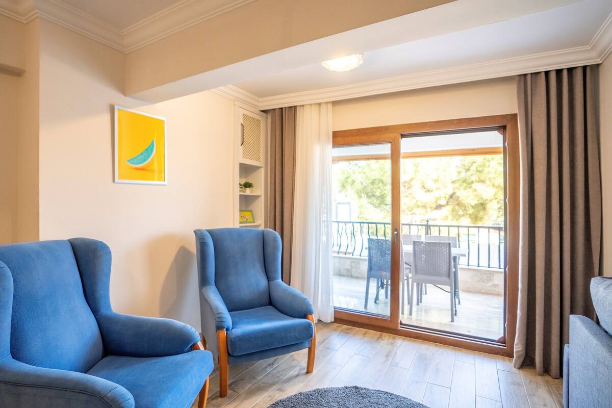 A living room with two blue armchairs, a grey rug, and a sliding glass door leading to an outdoor balcony with wicker furniture.