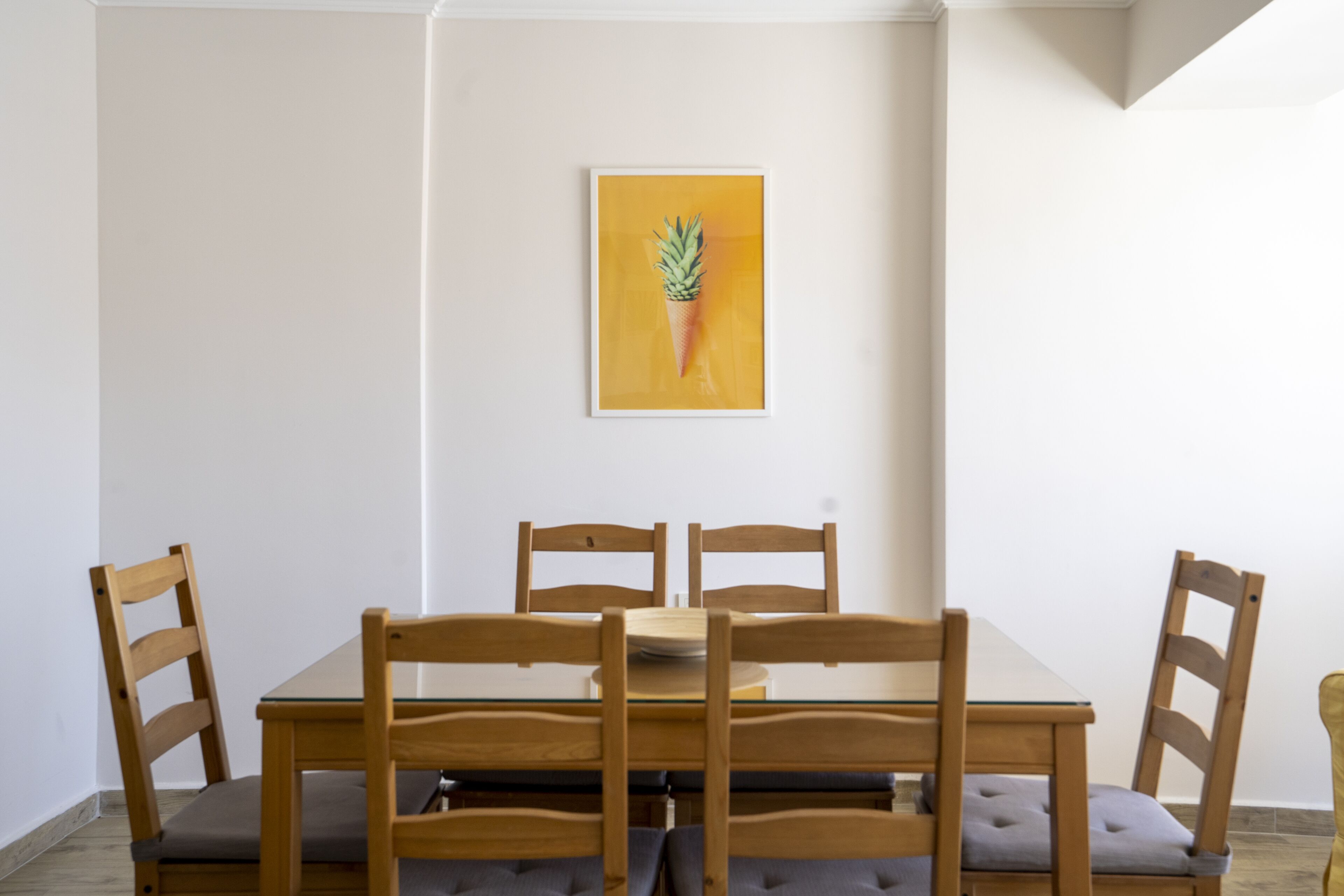 A dining area with a wooden table and four chairs, a framed picture on the wall, and a plain white wall.