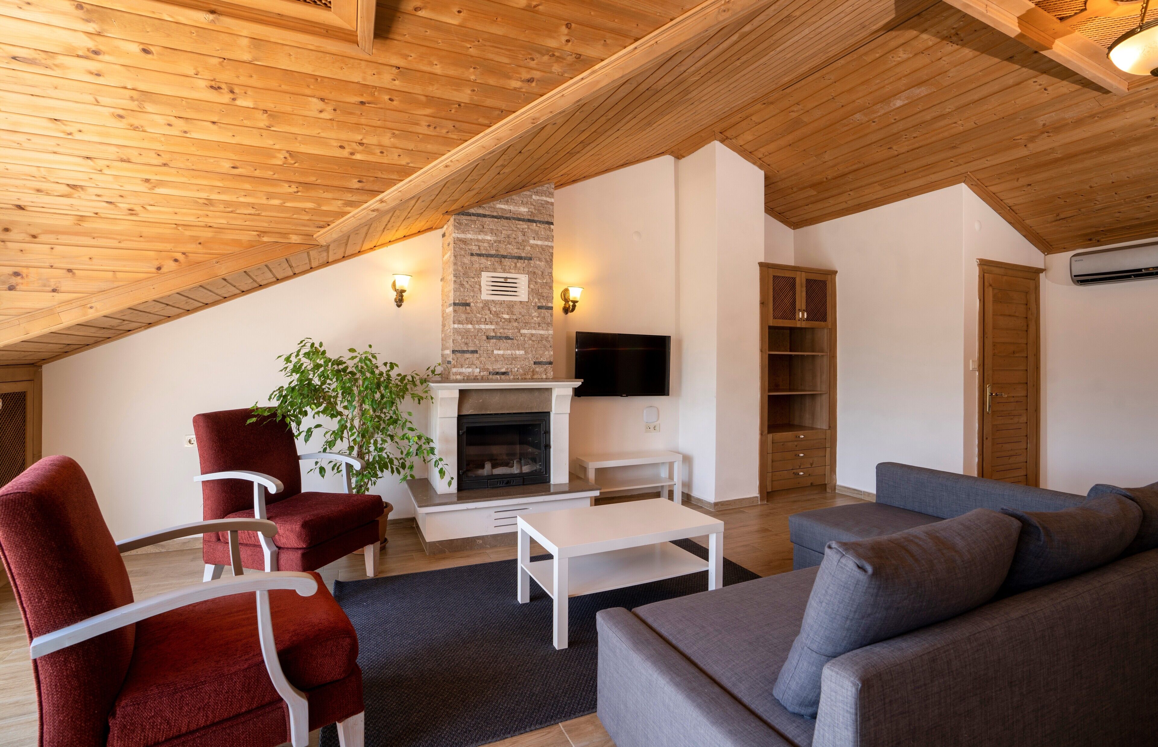 A cozy living room with a fireplace, red and grey sofas, a flat-screen TV, and wooden ceiling beams.