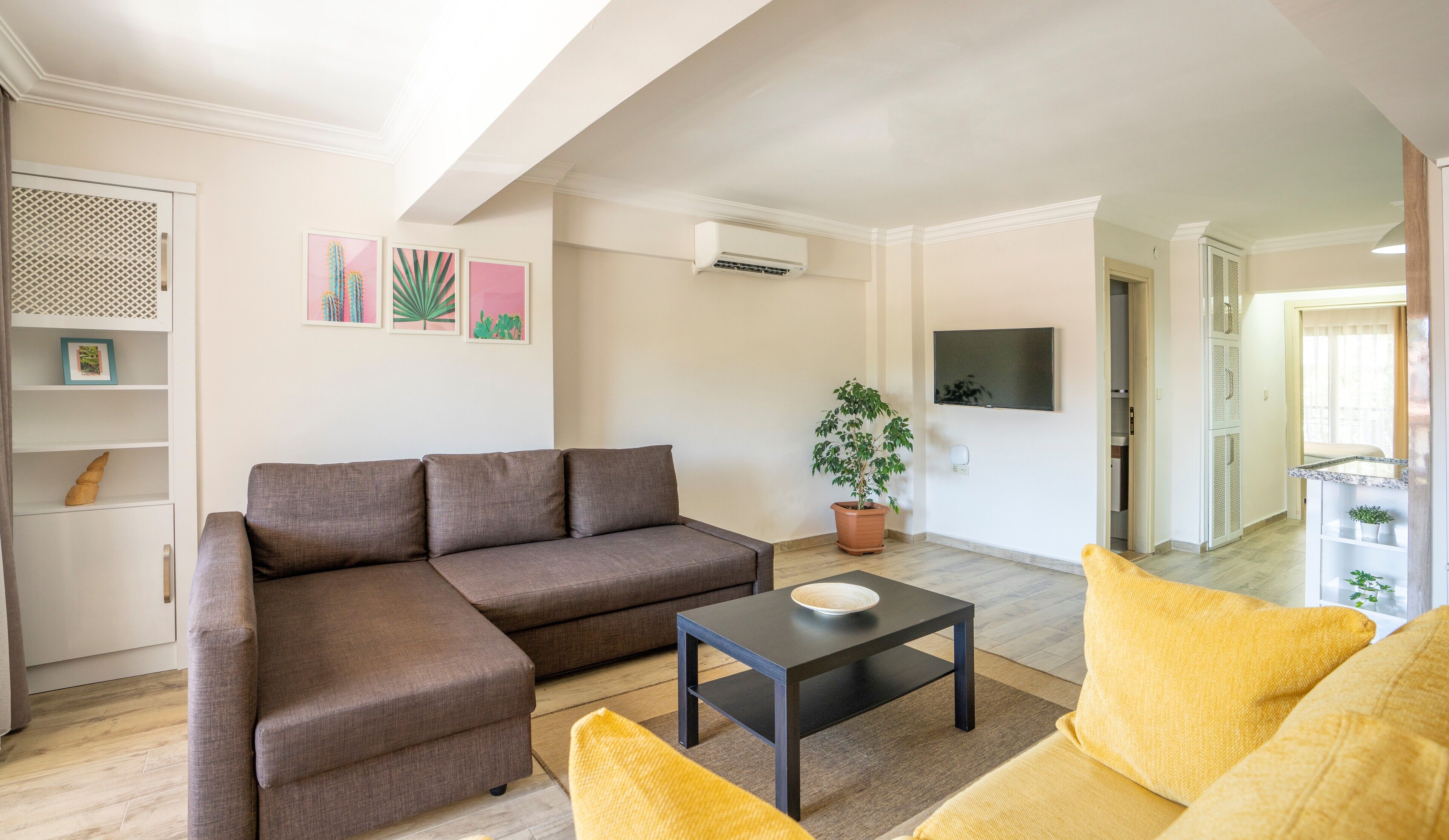 A living room with a brown sofa, a black coffee table, and a yellow armchair. There is a potted plant and a flat-screen TV mounted on the wall.