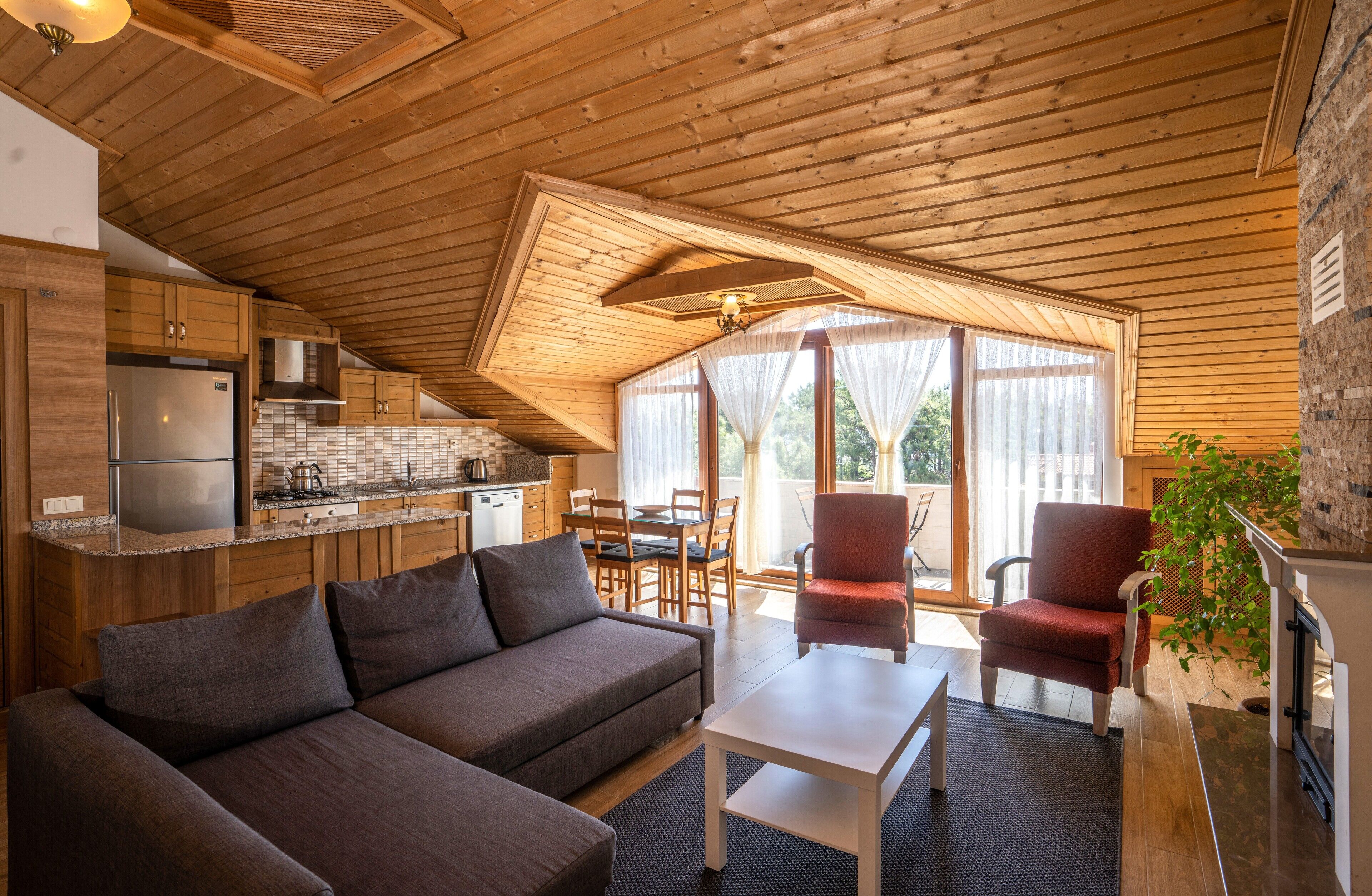 Een gezellige woonkamer met een bank, een eettafel en stoelen, een keuken met houten kastjes en een koelkast, en een groot raam met gordijnen.
