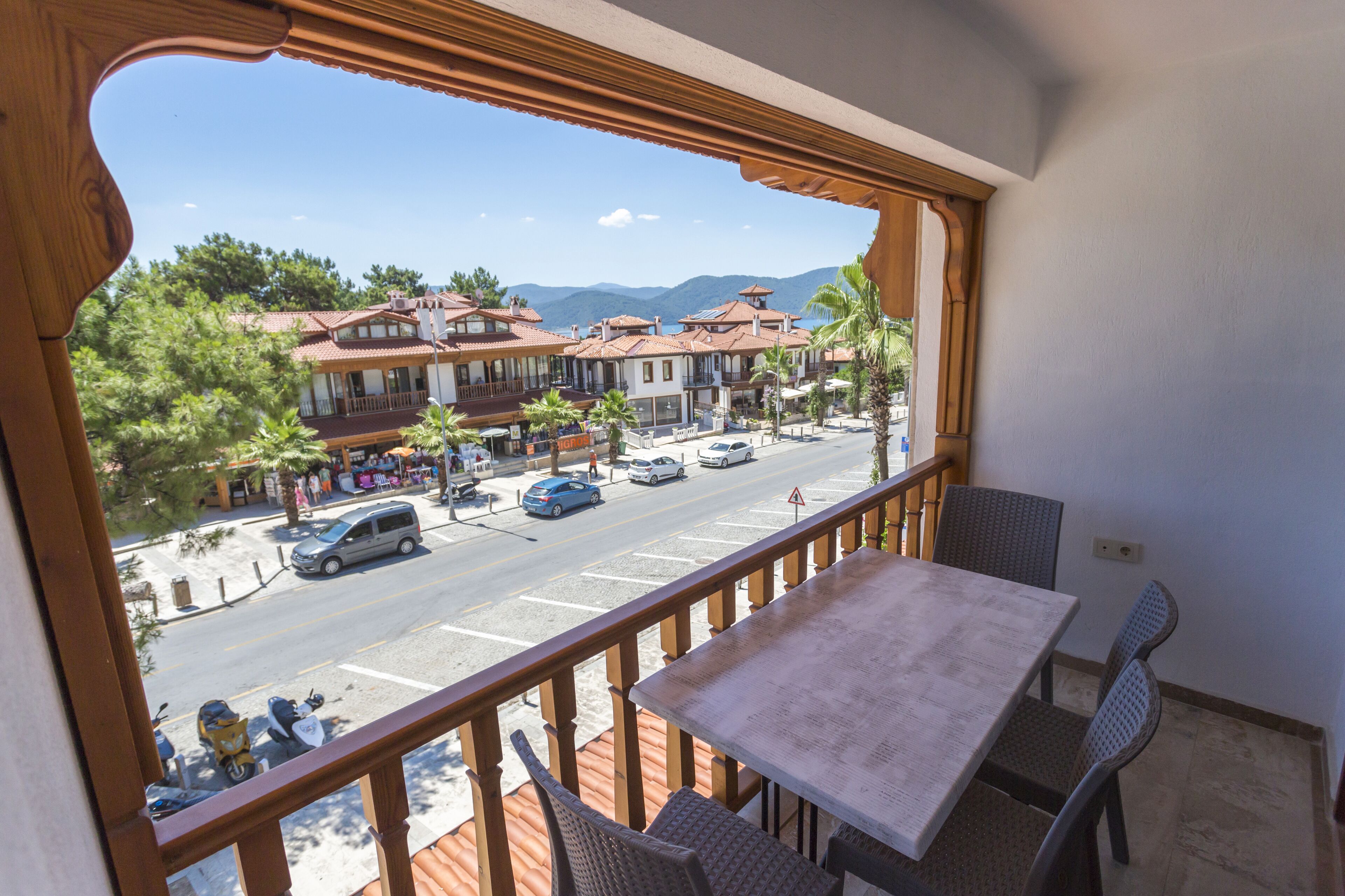 Un balcón con mesa y sillas, con vista a una calle con edificios y palmeras.