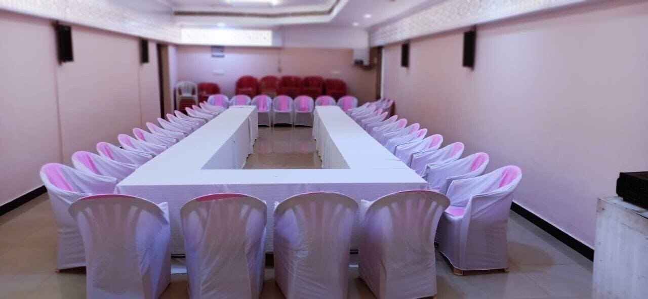 Un salón de conferencias con una larga mesa y filas de sillas, cubre-sillas rosas y sillas rojas al fondo.