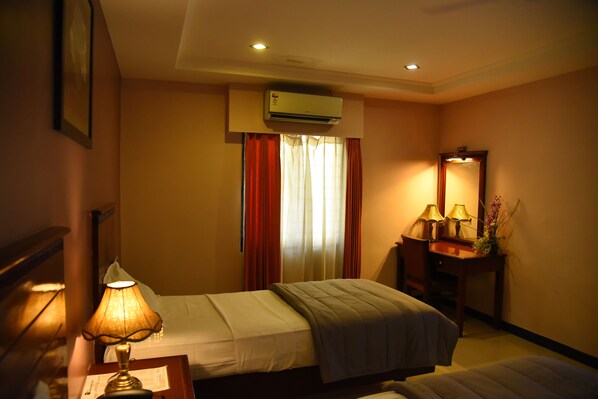 A hotel room with two beds, a bedside table with a lamp, a mirror, a vanity with a lamp, and an air conditioning unit above the window.