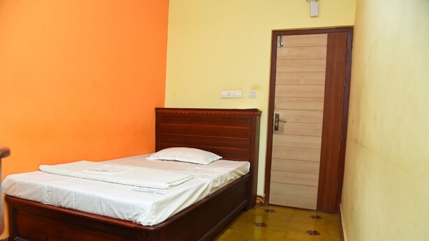 A single bed with a wooden headboard in a room with orange and yellow walls and a door with a wooden panel. - Hotel Subhalakshmi Palace (Karaikudi)