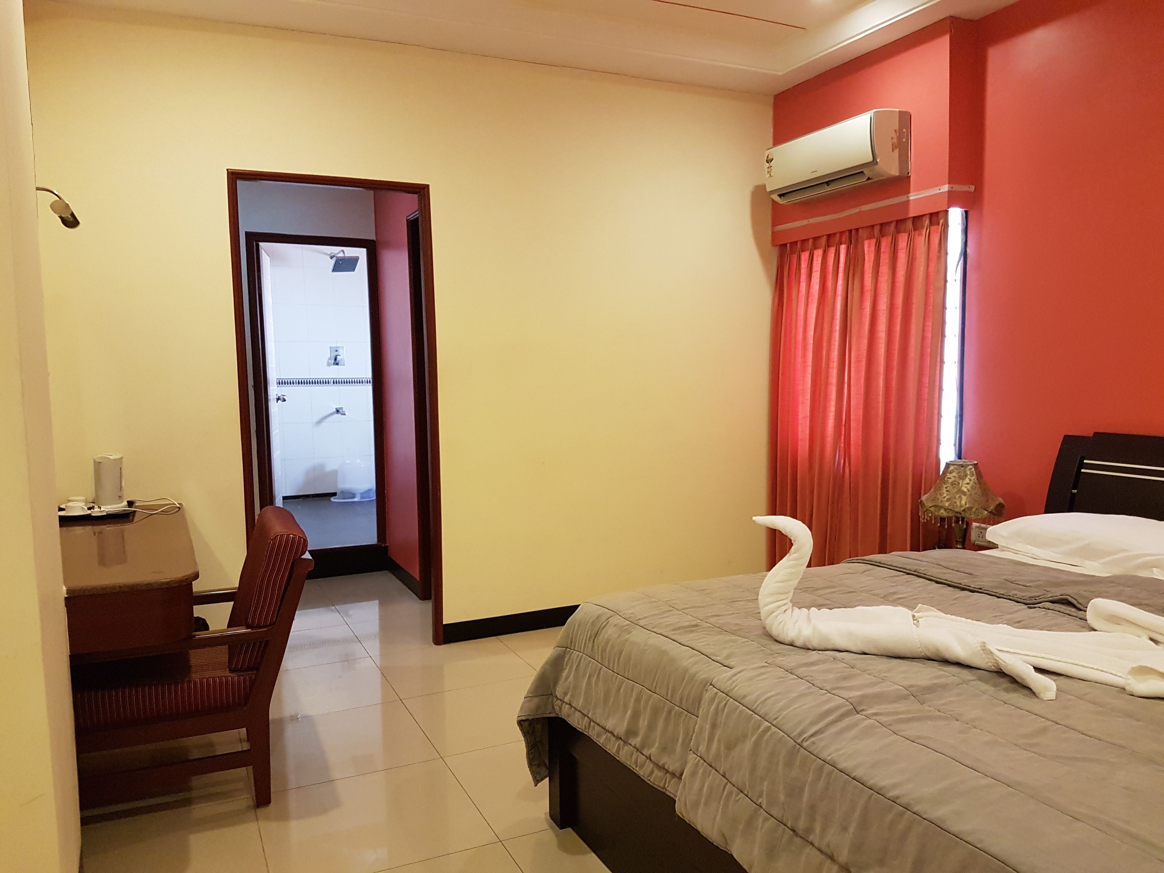 A hotel room with a bed, a desk, a chair, an air conditioning unit, and a view of the bathroom through an open door.