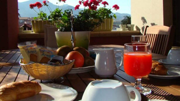 Una mesa de desayuno con vistas a montañas, una cesta de pan, una jarra de zumo de naranja y una taza de café.